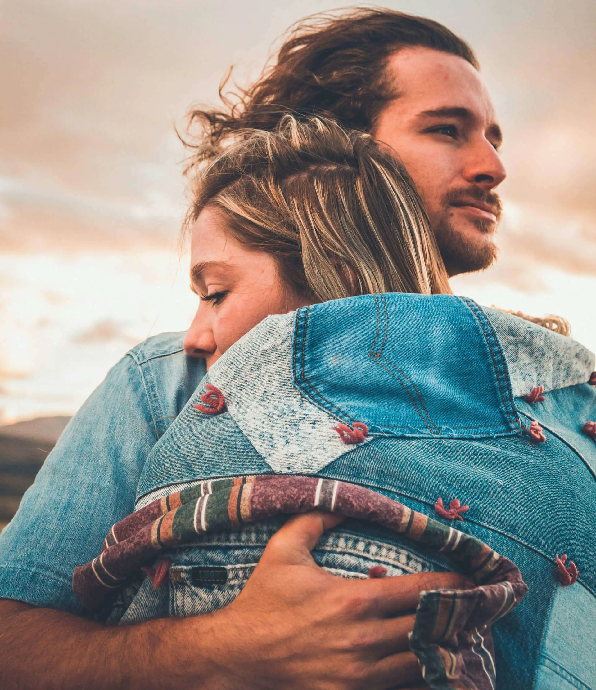 Man and woman embracing, both wearing denim jackets, with a serene sunset in the background