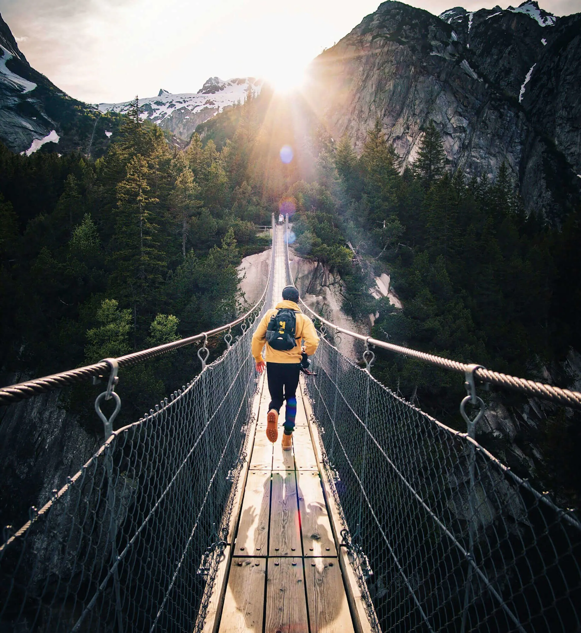 Person with a backpack walking on a suspension bridge in a mountainous area with the sun shining ahead