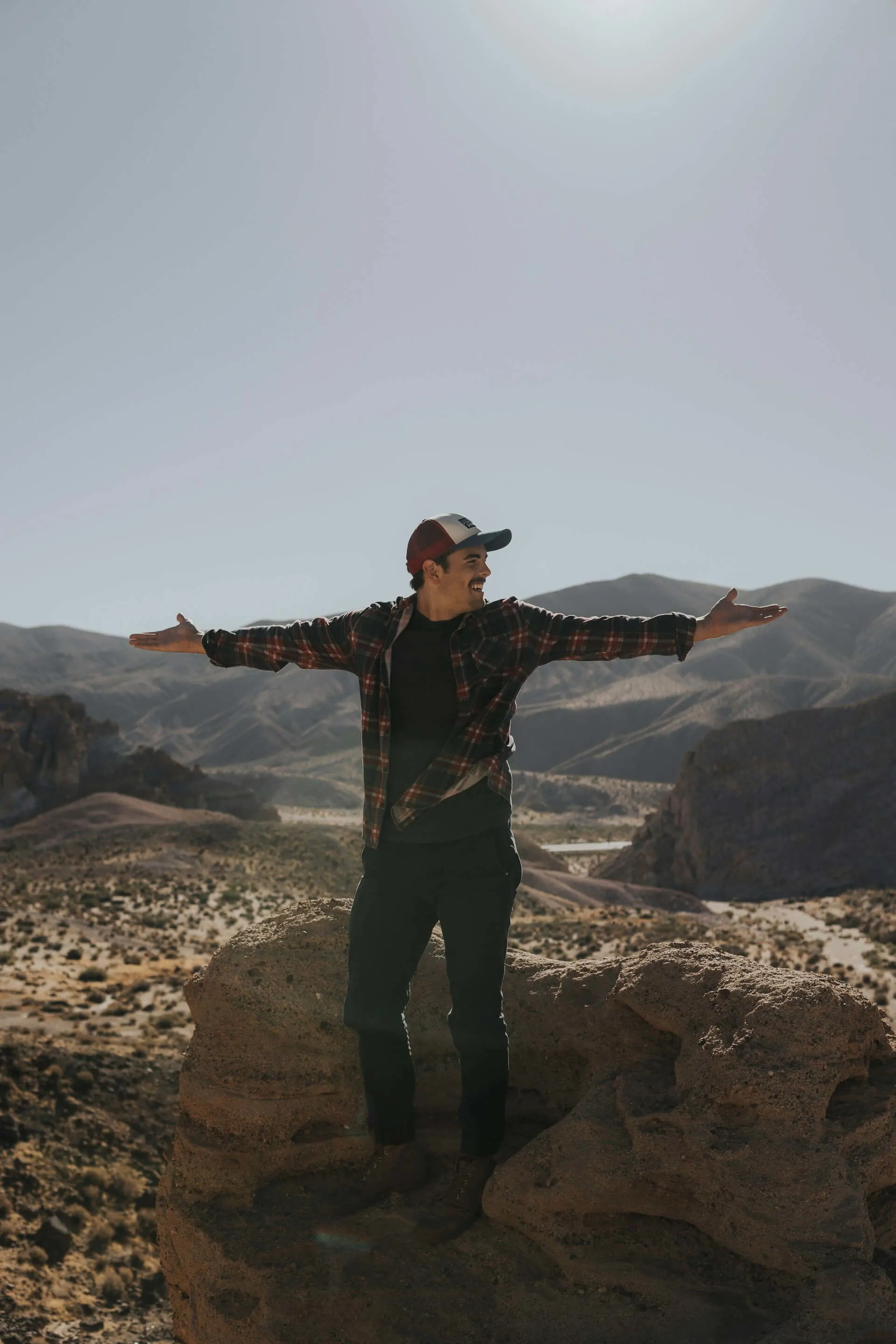 Man standing on a rock with arms outstretched, smiling, wearing a plaid shirt and a cap, with mountains in the background