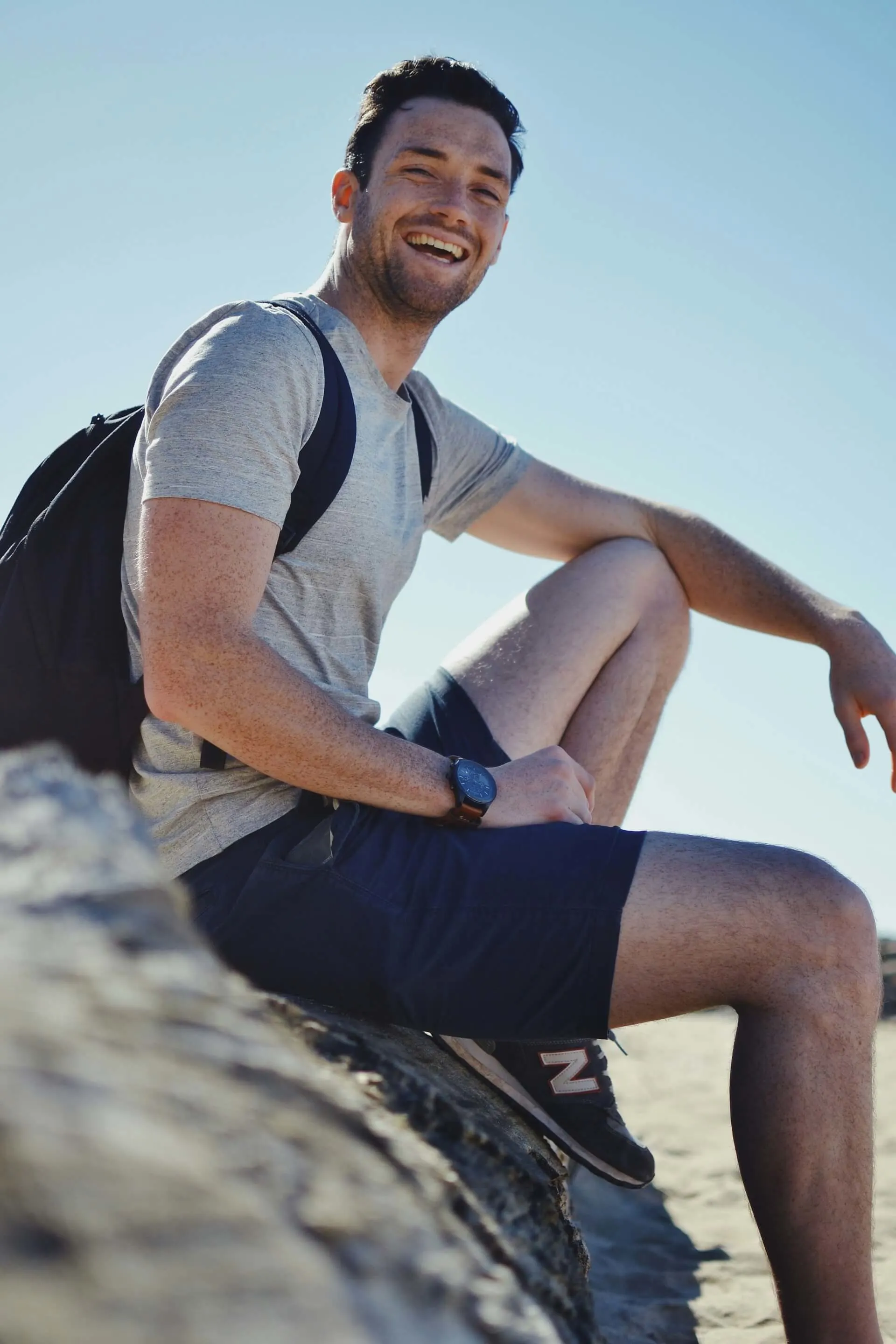 Smiling man sitting on a rock, wearing a backpack, with a bright blue sky in the background