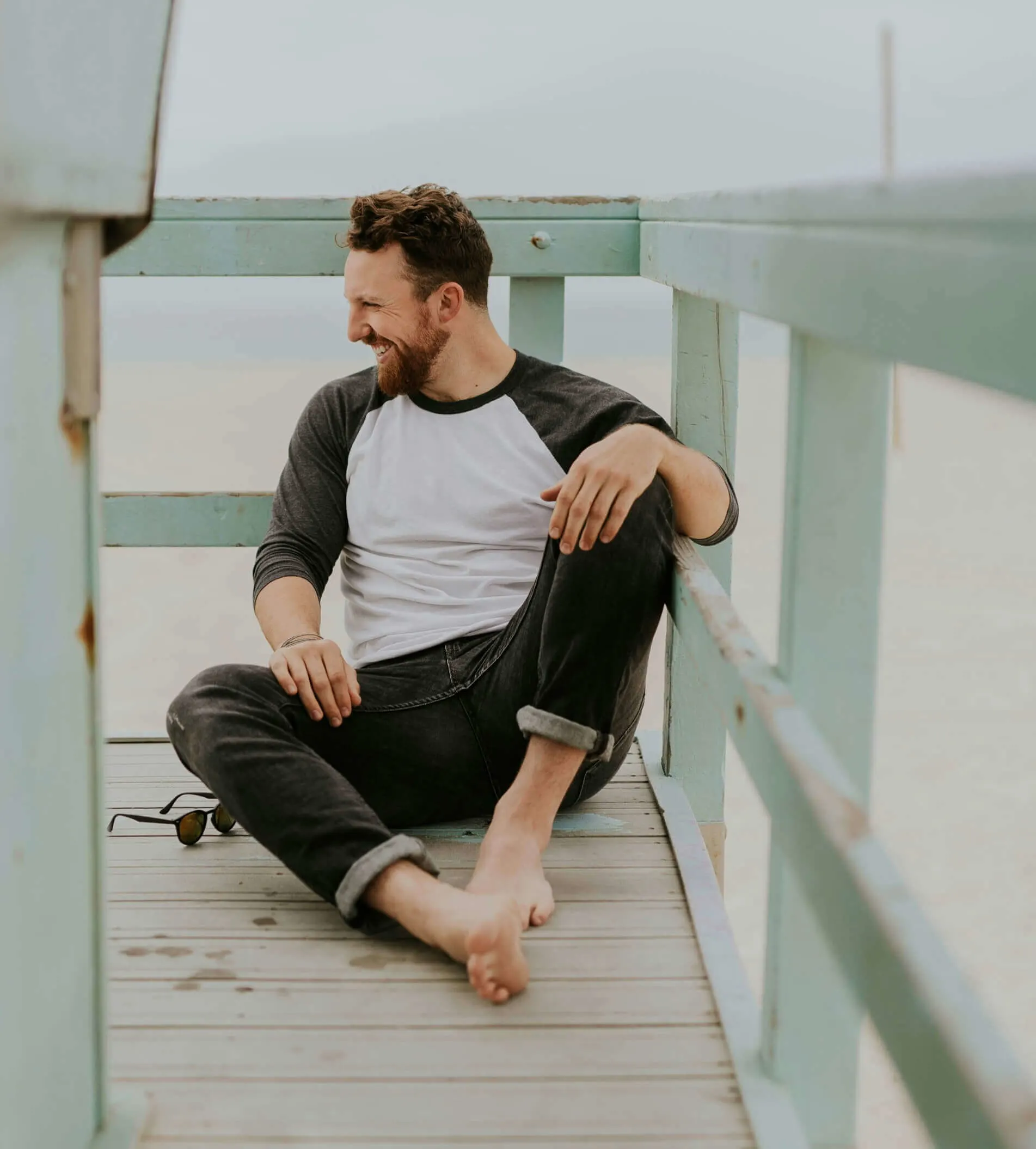 Man sitting barefoot on a lifeguard tower platform, smiling and looking to the side